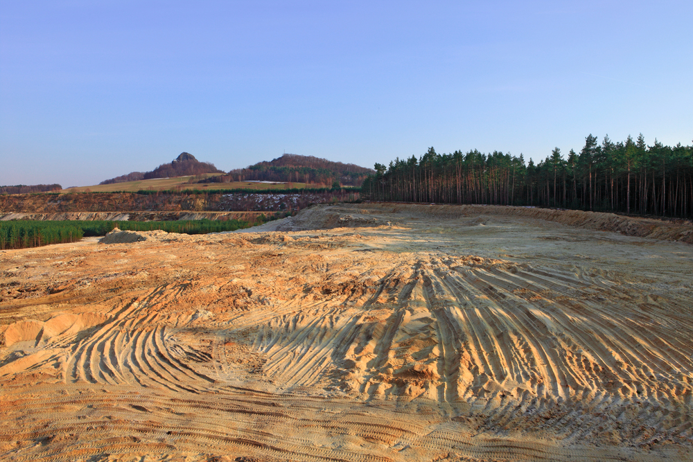 A wide expanse of sandy, cleared land with visible tire tracks, bordered by a forest and distant hills under a clear sky.