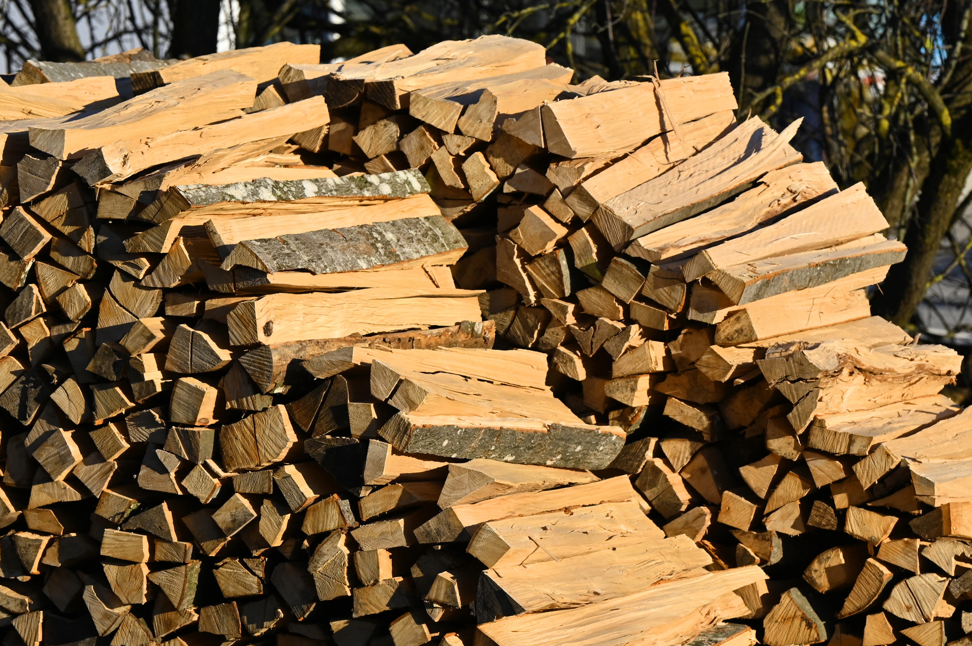 A pile of neatly stacked, split firewood logs outdoors, with some pieces showing bark and others bare wood.